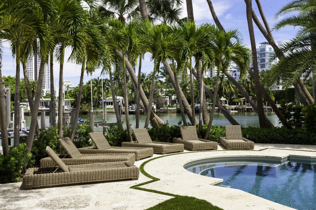Poolside scene with wicker lounge chairs and tall palm trees, overlooking a waterway. Nearby, modern buildings rise against a clear sky.