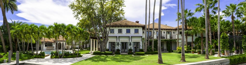 Luxurious mansion with terracotta roof, surrounded by palm trees and well-manicured lawns, under a clear blue sky, conveying tranquility and elegance.