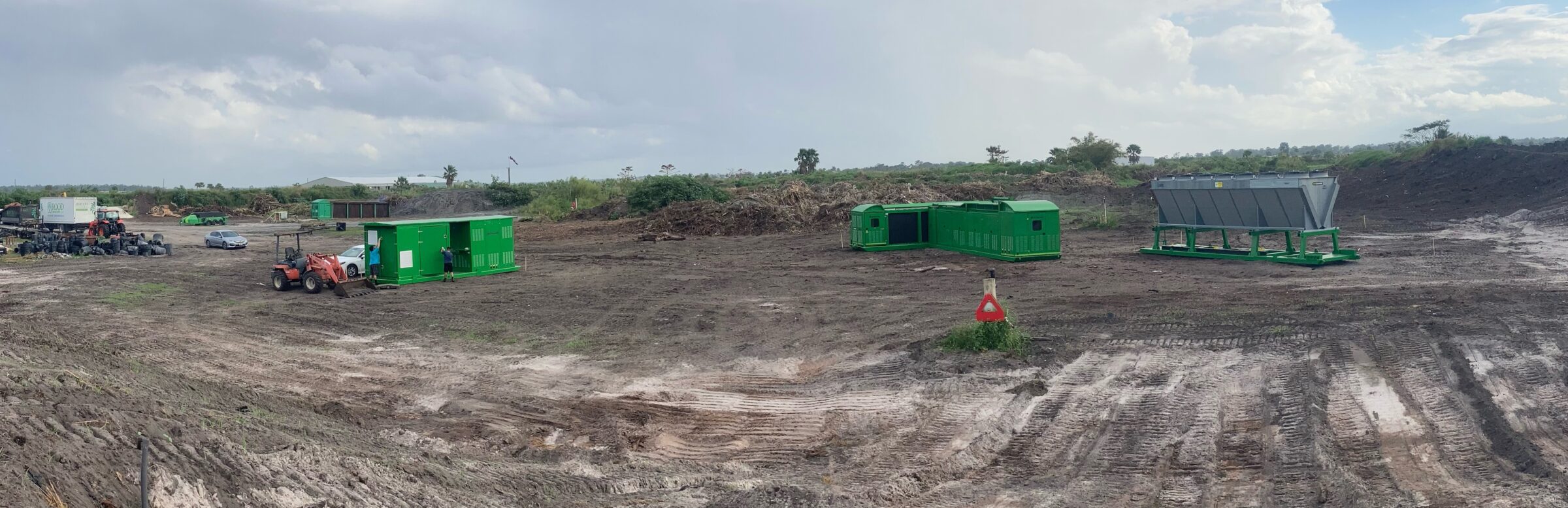 A person near green containers and a tractor on a large, cleared construction site with piles of tires and cloudy sky.