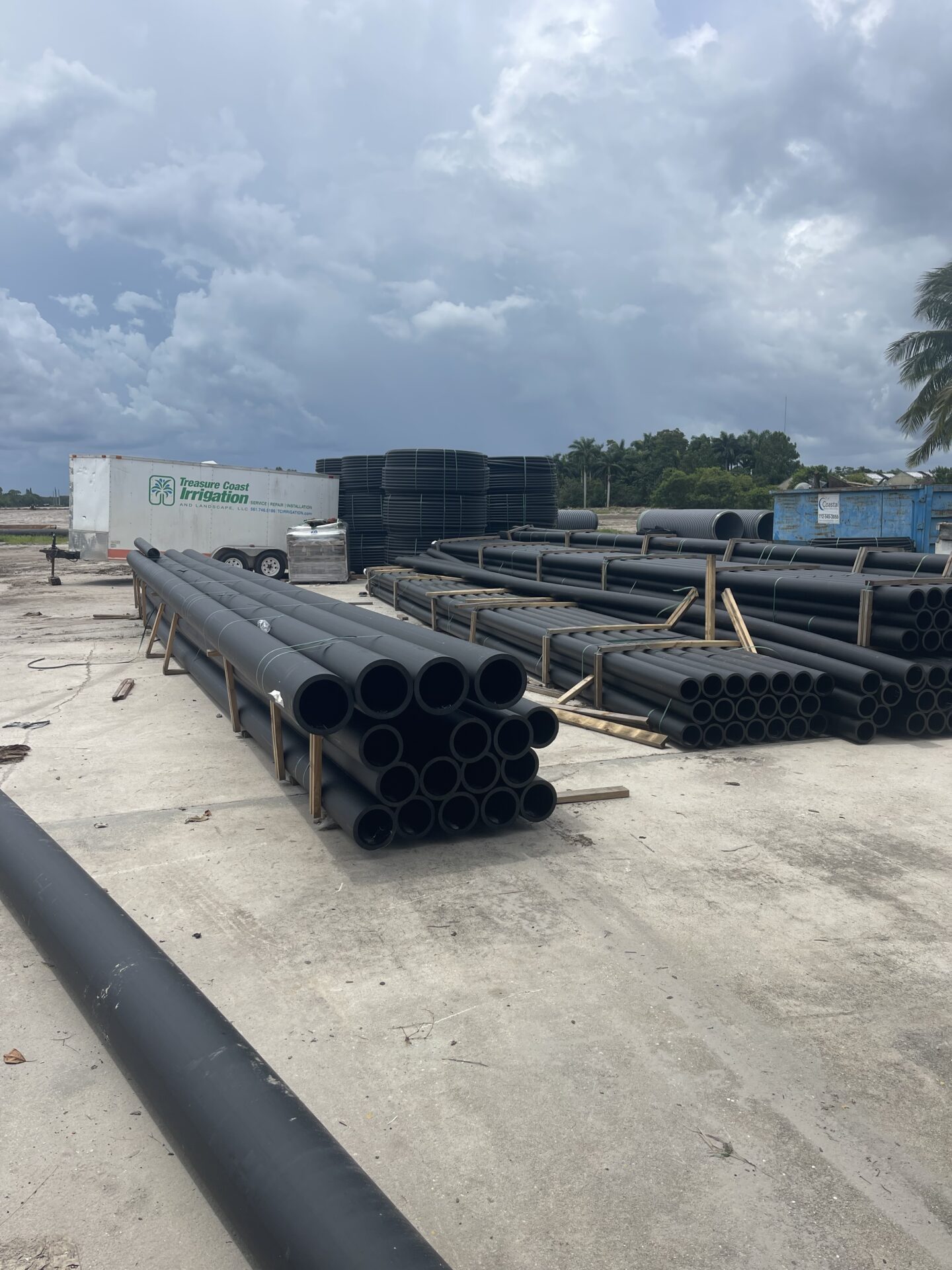 Construction site with stacked black pipes, a trailer labeled "Treasure Coast Irrigation," and cloudy sky. Palm trees line the background.