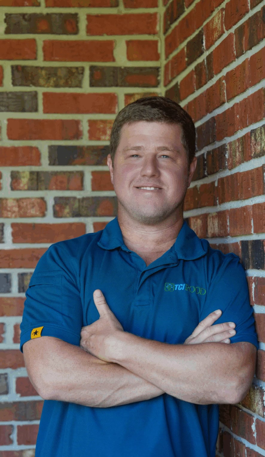 Person in a blue shirt confidently stands against a red brick wall backdrop, smiling with arms crossed, creating a casual and approachable impression.