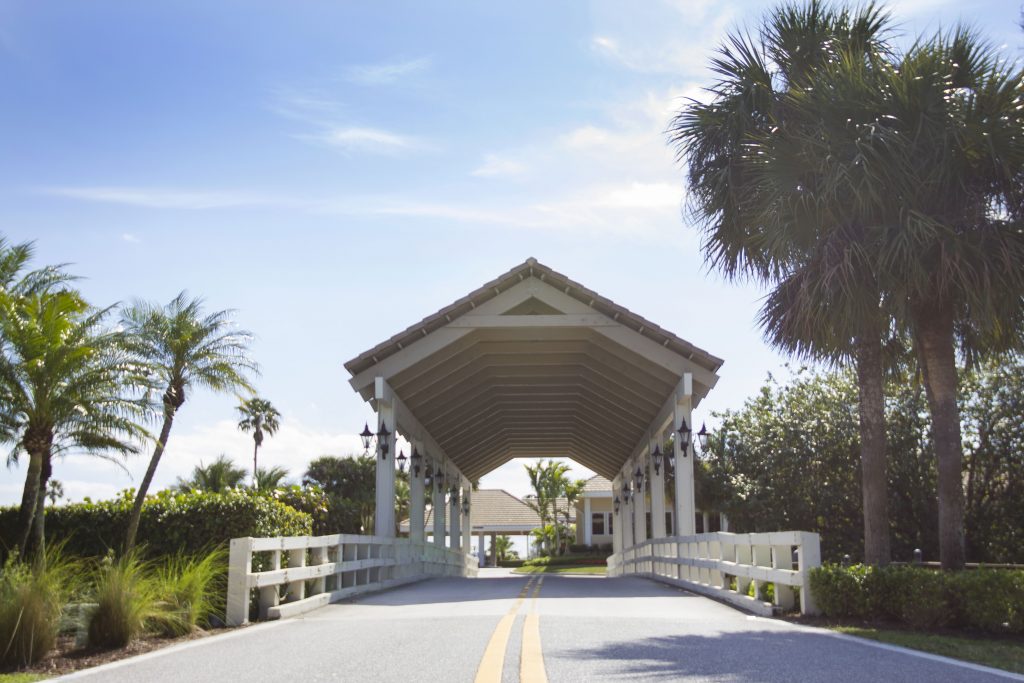 A white covered bridge over a paved road, surrounded by palm trees under a clear blue sky. No recognizable landmarks or historical buildings.
