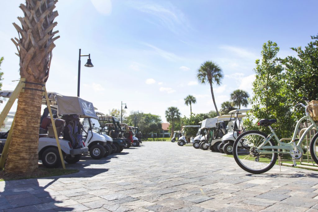 A row of golf carts and a bicycle line a sunny, palm-lined street, surrounded by lush greenery and a clear blue sky.