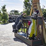 A row of golf bags is neatly placed on a stand beside parked golf carts, surrounded by trees and a paved path.
