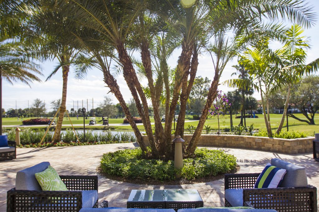 Lush patio with wicker furniture, cushions, and palm trees overlooking a golf course. Manicured green and serene environment under clear skies.