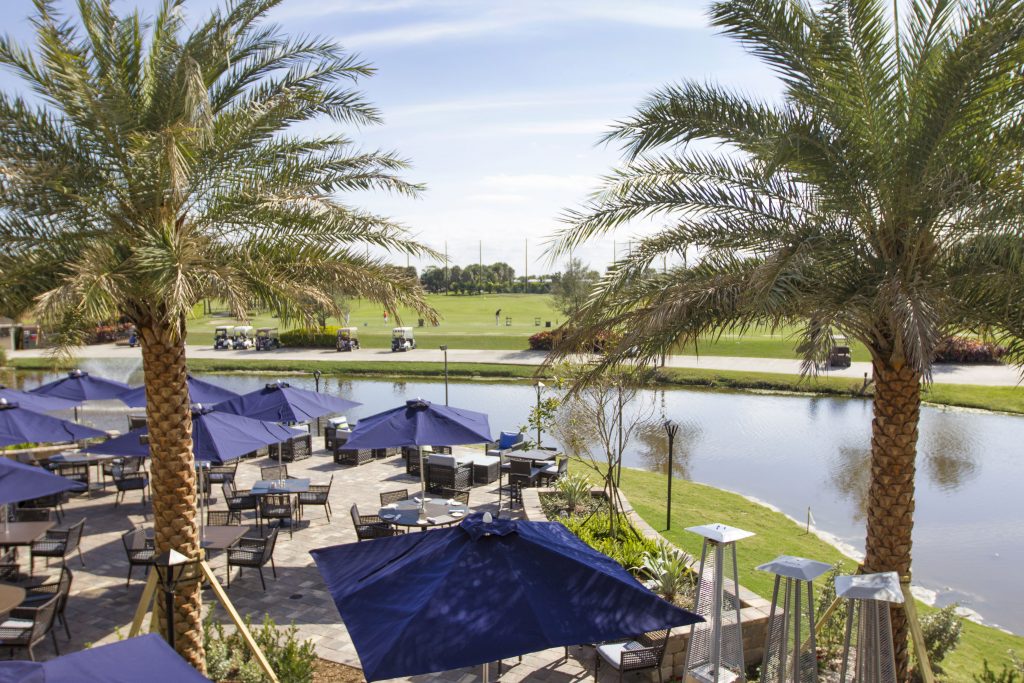 A golf course with outdoor dining area featuring blue umbrellas, palm trees, and a pond. Golf carts and people are visible in the distance.