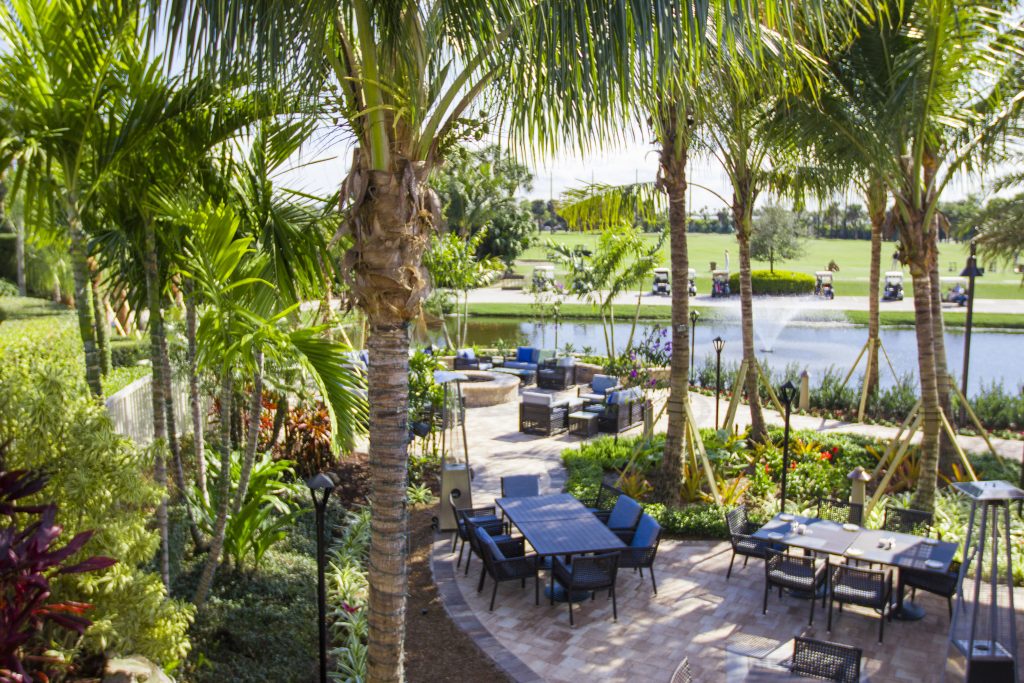 Tropical landscape with palm trees, patio furniture, and a pond with a fountain. Golf carts are visible on the green field beyond.