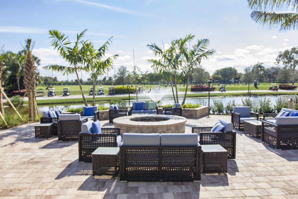Outdoor patio with wicker seating and blue cushions around a fire pit. In the background, golf carts and a fountain are visible.