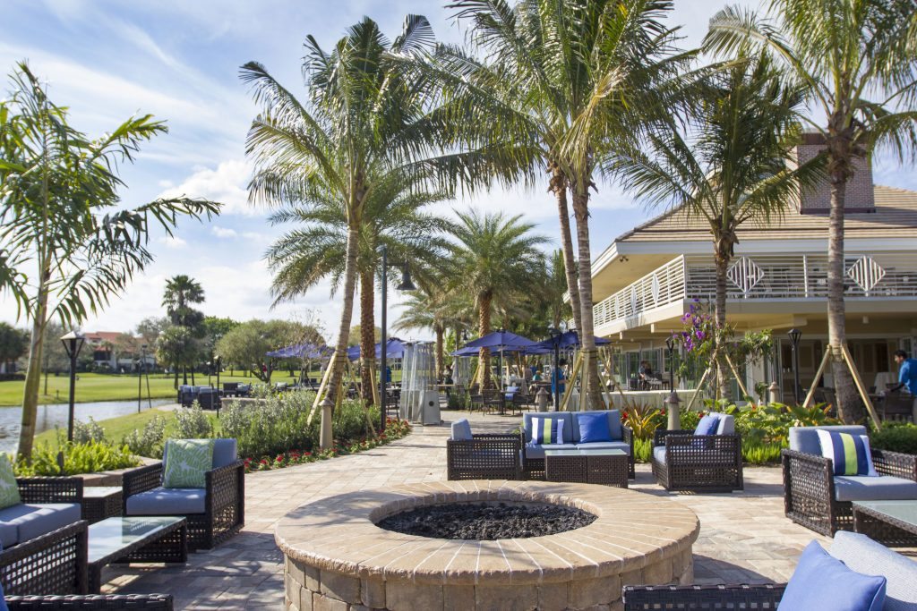 Outdoor seating area with palm trees, fire pit, and a building with balconies. Bright, sunny weather; blue cushions and umbrellas enliven the scene.