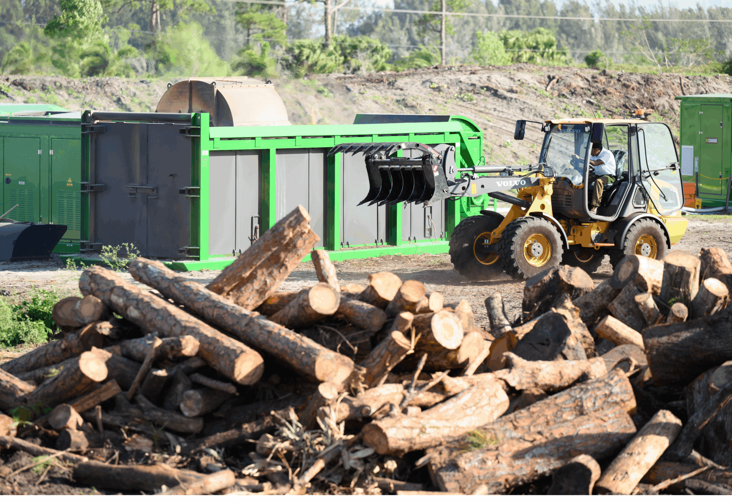 A person operates a loader near a wood pile and industrial machinery on a construction site, surrounded by trees and greenery.