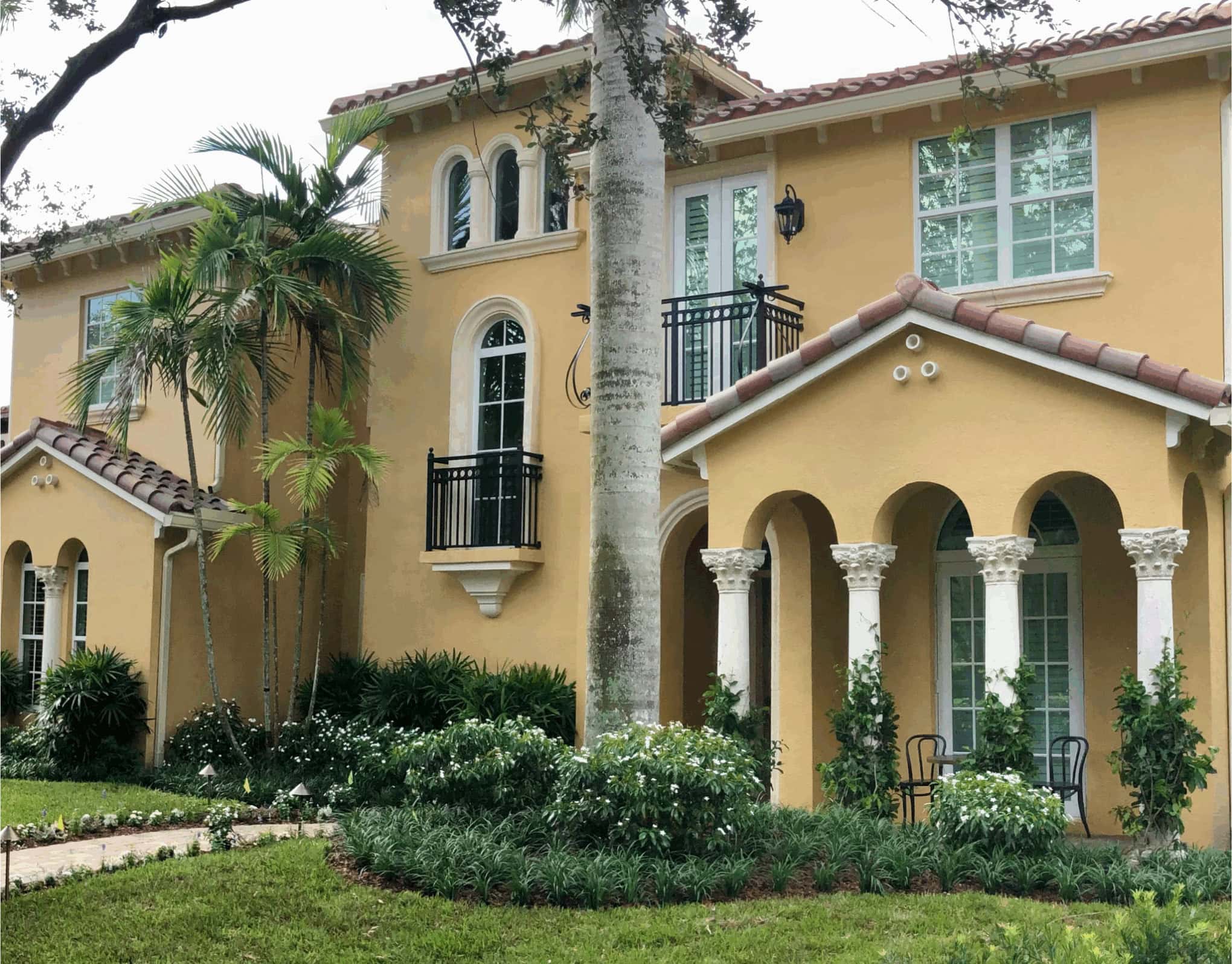 Elegant yellow house with arched columns, tiled roof, and lush greenery. A small seating area enhances the inviting, Mediterranean-style architecture.