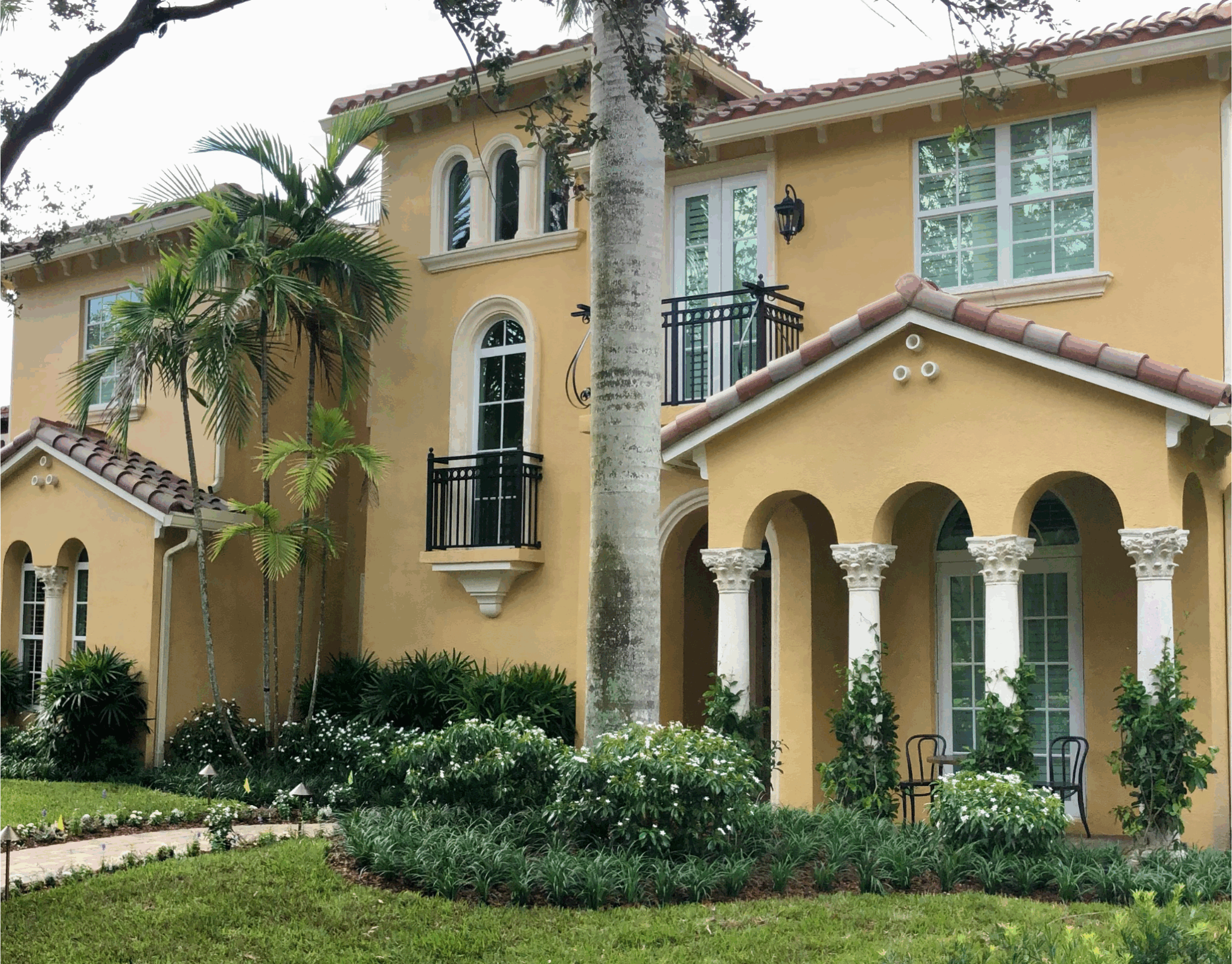 Elegant yellow house with arched columns, tiled roof, and lush greenery. A small seating area enhances the inviting, Mediterranean-style architecture.